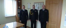 Na zdjęciu w biurze komendanta powiatowego stoją trzej strażacy ubrani w czarne mundury wyjściowe (gabardyna)