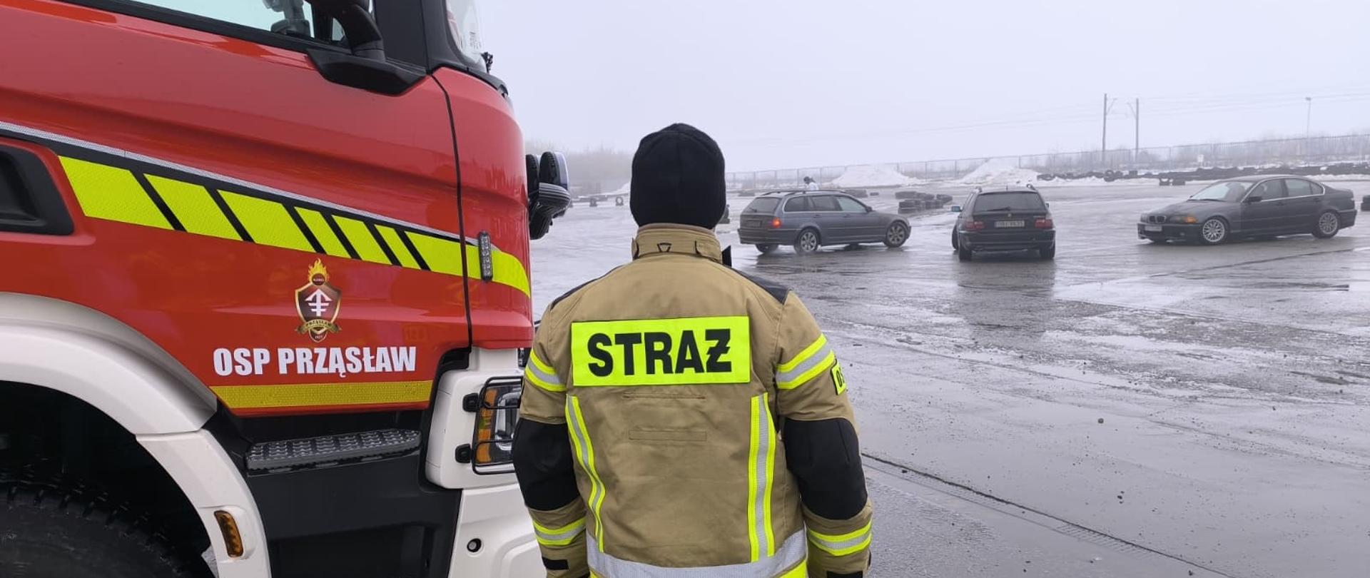 Na zdjęciu lewy przód samochodu ratowniczo-gaśniczego. Obok strażak odwrócony plecami do fotografa. Strażak patrzy w stronę toru do pokazu dryftu i off-road’u.