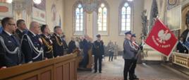 Policjanci stoją ze sztandarem, w ławkach stoją przedstawiciele służb mundurowych oraz zaproszonych gości.