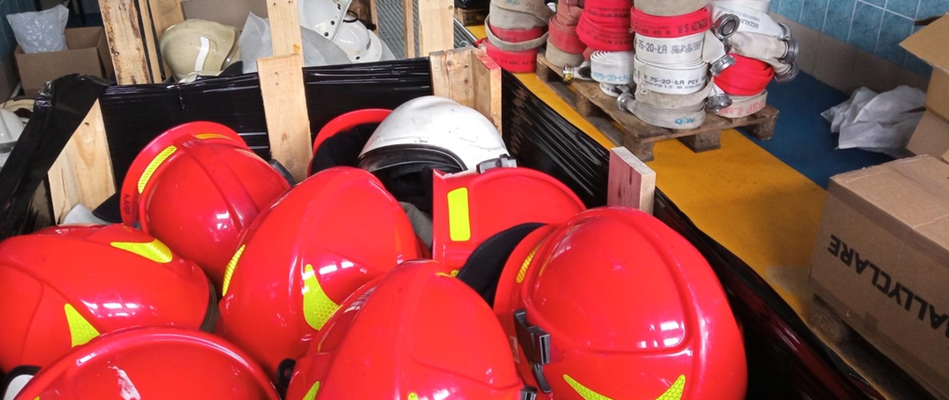 Na zdjęciu widoczne hełmy strażackie koloru czerwonego i białego w oddali węże pożarnicze spakowane na paletach przygotowane do transportu dla strażaków z Ukrainy.