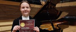 Dziewczynka stoi na estradzie sali koncertowej przed fortepianem i trzyma w rękach dyplom.