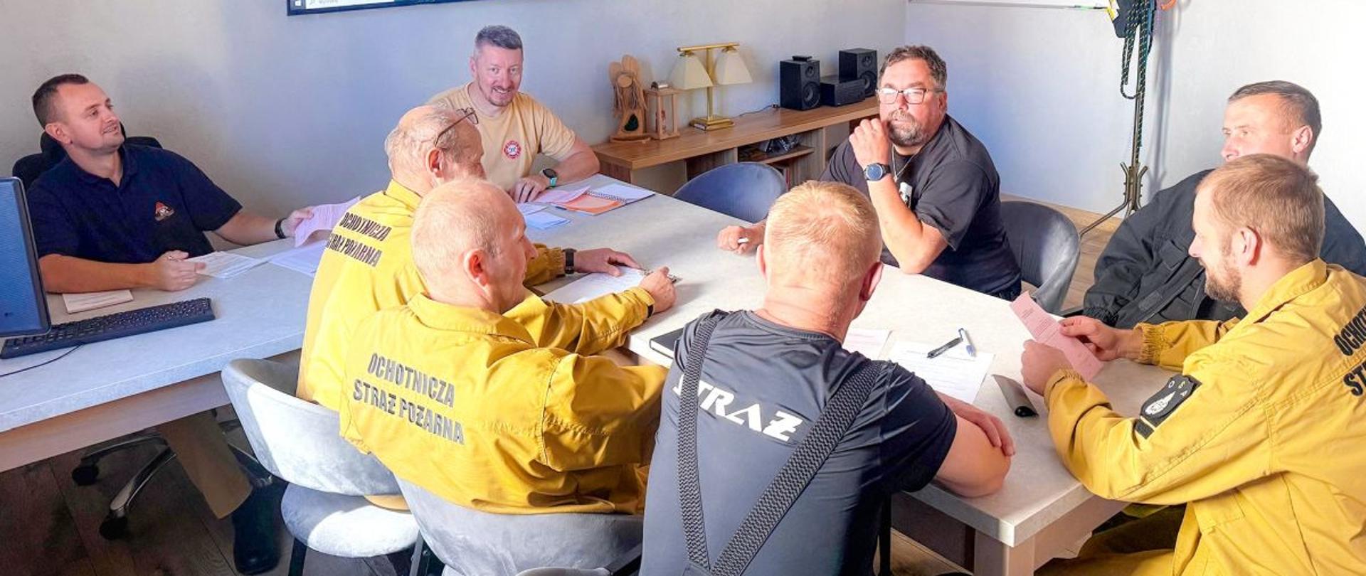 Zdjęcie wykonane jest na świetlicy JRG w Ustrzykach Dolnych. Przy dwóch stołach ustawionych do siebie prostopadle siedzą strażacy OSP biorący udział w szkoleniu KDRa. Z lewej strony przy komputerze siedzi strażak PSP który prowadzi szkolenie. Na środku u góry zdjęcia na ścianie zawieszony jest telewizor na którym wyświetlany jest materiał szkoleniowy. 