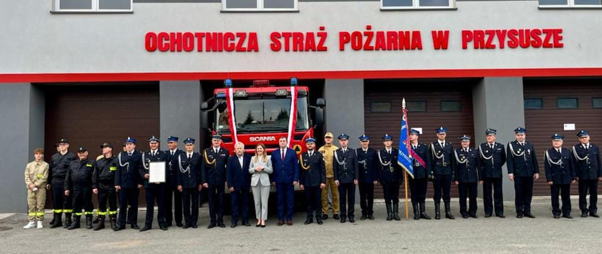 Zdjęcie przedstawia dużą grupę osób, głównie strażaków w galowych mundurach, stojących przed remizą Ochotniczej Straży Pożarnej w Przysusze. Budynek remizy jest szary z dużym, czerwonym napisem "OCHOTNICZA STRAŻ POŻARNA W PRZYSUSZE" nad trzema bramami garażowymi. W centralnej części grupy, przed środkową bramą, stoi czerwony wóz strażacki marki Scania, udekorowany biało-czerwonymi wstążkami. Obok wozu i przed nim, stoi w szeregu około dwudziestu kilku osób. Większość z nich to strażacy w granatowych mundurach galowych i rogatywkach. W środku grupy stoją również dwie osoby w strojach cywilnych: kobieta w jasnym garniturze i mężczyzna w ciemnym garniturze, prawdopodobnie przedstawiciele władz lokalnych lub zaproszeni goście. Po prawej stronie grupy strażaków jeden z nich trzyma sztandar. Cała scena sugeruje uroczystość związaną z przekazaniem wozu strażackiego lub inną ważną ceremonię dla jednostki OSP w Przysusze. Jest to dzień z pochmurnym niebem.