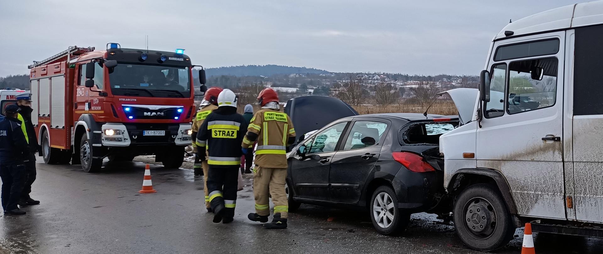 Fotografia przedstawia białego busa, który uderzył w tył czarnego samochodu osobowego. Za nimi znajduje się samochód Straży Pożarnej na sygnałach. Pomiędzy samochodami stoją strażacy oraz policjanci.