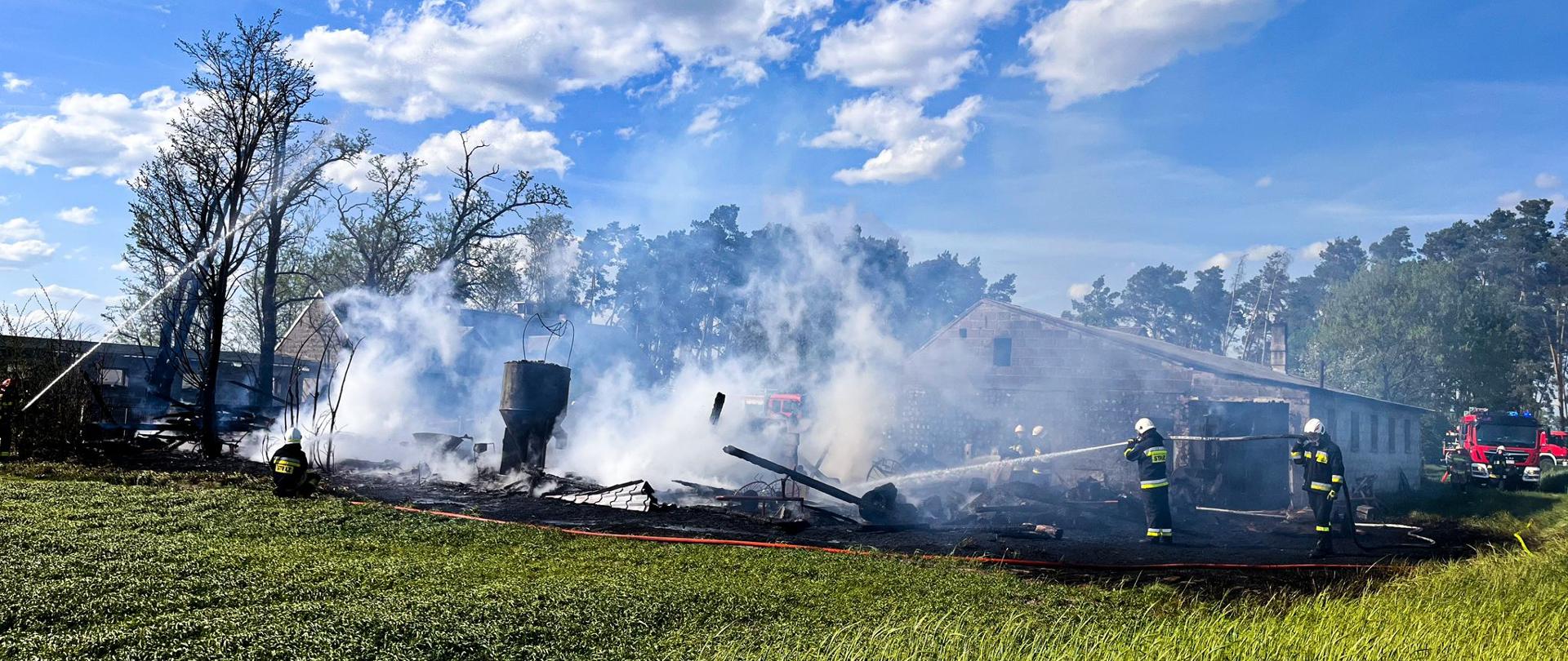 Strażacy gaszą pozostałości spalonego budynku. W tle widać obroniony obiekt i wóz strażacki.