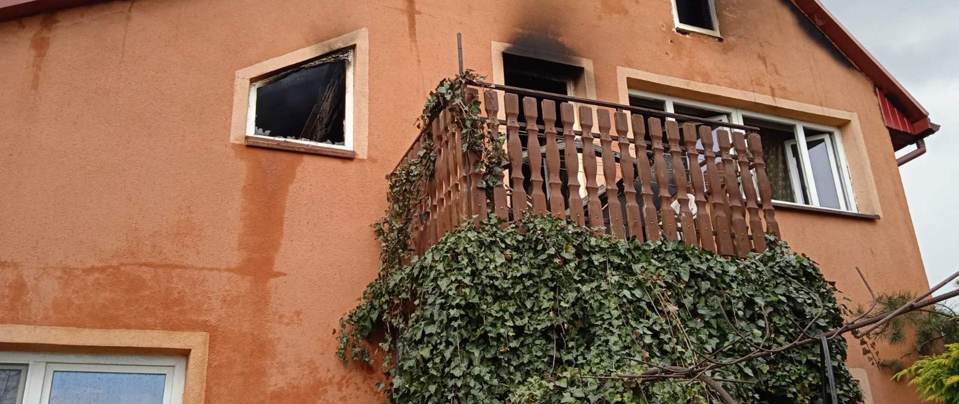 Ściana frontowa domu mieszkalnego, elewacja koloru pomarańczowego, w ścianie widoczne otwory okienne, przy których występują okopcenia, na wyższej kondygnacji widoczny balkon. Przed wejściem do budynku stoją strażacy. 