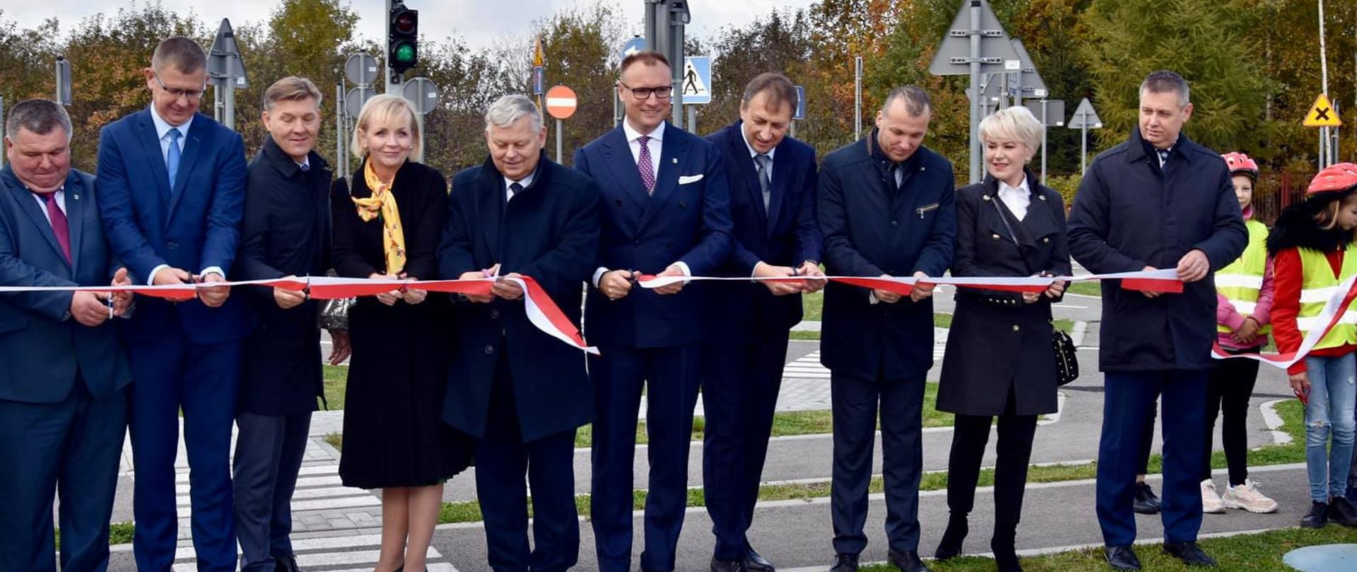 Zdjęcie przedstawia moment symbolicznego przecięcia biało-czerwonej wstęgi. Na zdjęciu znajdują się elegancko ubrane osoby. 