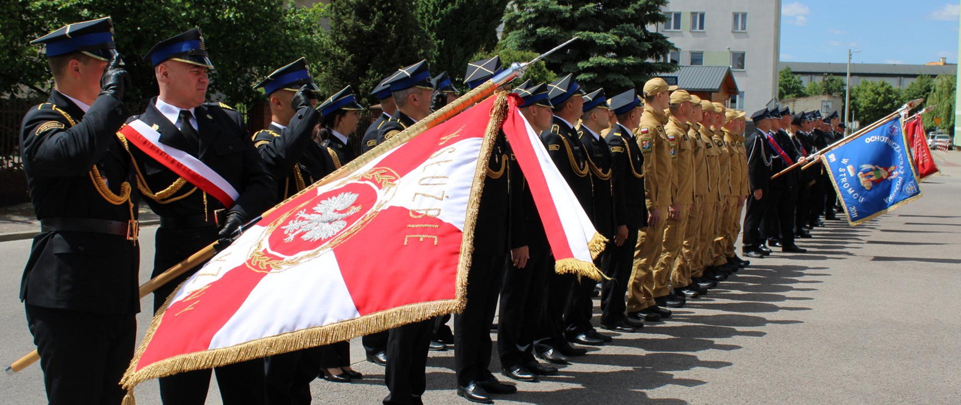 Uroczysty apel strażacki na ulicy w Białobrzegach. Wytwornie umundurowani strażacy stoją w dwuszeregu, trzymając sztandary jednostek. Pierwsi strażacy z pocztu sztandarowego salutują. W tle widoczne są bloki mieszkalne i drzewa.