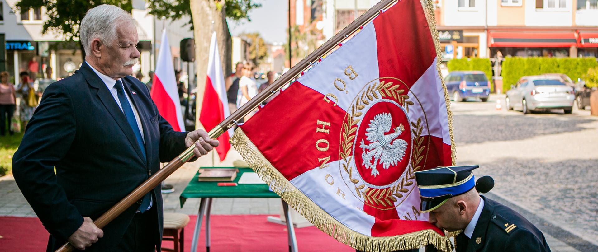 Wicewojewoda Pomorski Pan Mariusz Łuczyk trzymając pochylony sztandar wręcza go Komendantowi Powiatowemu Państwowej Straży Pożarnej w Lęborku. Komendant Powiatowy Państwowej Straży Pożarnej w Lęborku mł. bryg. Piotr Krzemiński klęcząc całuje płat odbieranego sztandaru. 