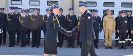 Przywitanie Zastępcy Komendanta Wojewódzkiego Państwowej Straży Pożarnej w Łodzi przez Dowódcę uroczystości. W tle widać strażaków ustawianych w dwuszeregu ubranych w umundurowanie galowe i koszarowe.