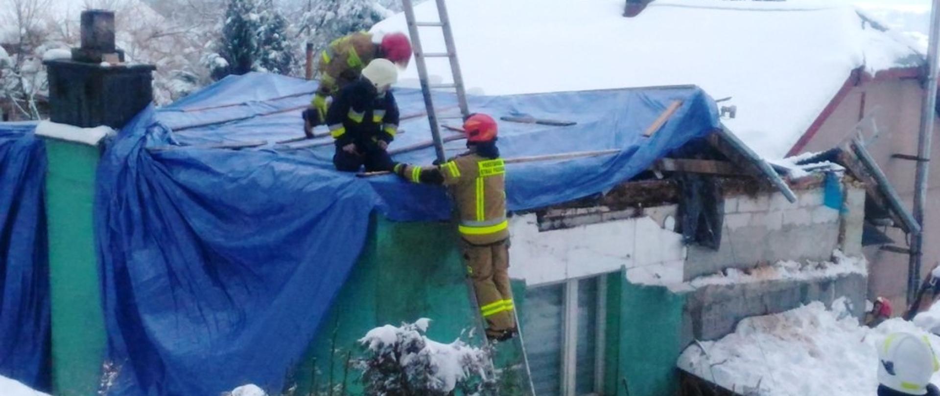 Kolorowa fotografia przedstawia na pierwszym planie część budynku z zawalonym dachem, przykrytego doraźnie niebieskimi plandekami. Na dachu dwaj umundurowani strażacy zabezpieczają listwami plandeki. Obok nich na drabinie opartej do ścianę budynku stoi i pomaga trzeci strażak. W tle widać inne mocno zaśnieżone budynki i innych strażaków w akcji.