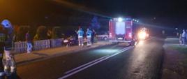 Widok z oddali: samochód z uszkodzonym przodem, stoi przy płocie z podniesioną maską. Obok stoją strażacy z PSP i samochody pożarnicze.