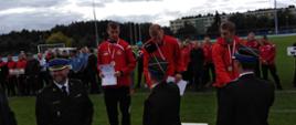 Trzech mężczyzn w strojach sportowych stoi na podium. Przed nimi stoi trzech strażaków. W tle widać sportowców , drzewa i duży budynek. 
