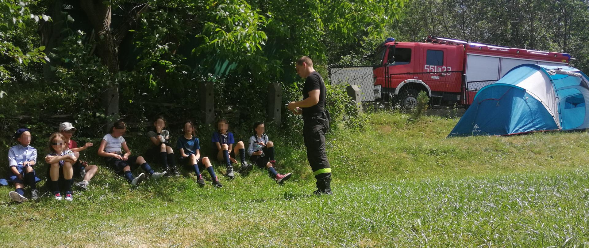 Widok z boku. Strażak PSP stoi przed harcerzami, którzy siedzą na trawie i prowadzi prelekcję. Na drugim planie niebieski namiot oraz samochód ratowniczo-gaśniczy PSP.
