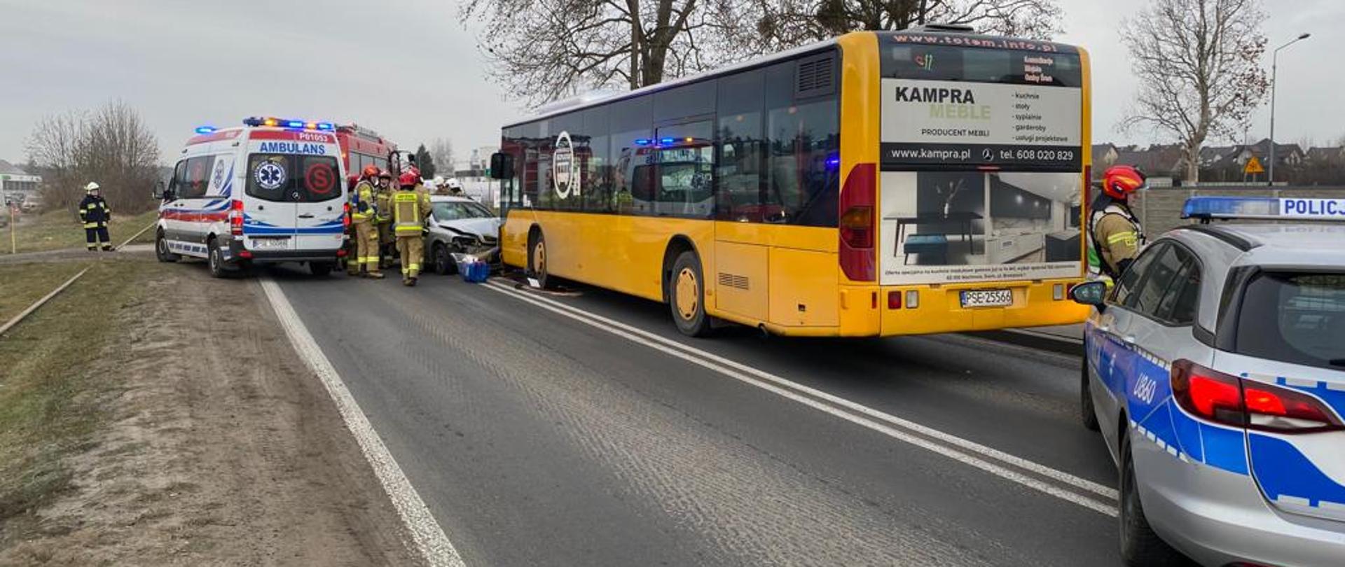 Na zdjęciu autobus koloru żółtego oraz samochód osobowy koloru srebrnego zderzone razem czołowo , przed autobusem samochód Policji, obok samochodu osobowego karetka, strażacy w czerwonych i białych hełmach stoją przy rozbitych autach