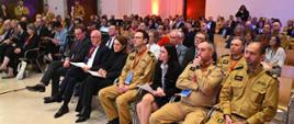 Grupa osób siedzących na sali konferencyjnej. Wśród nich są straży ubrani w mundury 