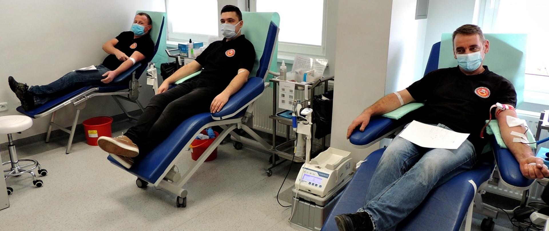 Na zdjęciu trzech strażaków w czarnych koszulkach i dżinsach siedzą na granatowych fotelach oddają krew w pomieszczeniu o białych ścianach i szarej posadzce w Regionalnym Centrum Krwiodawstwa i Krwiolecznictwa w Oddziale Terenowym w Gorlicach.