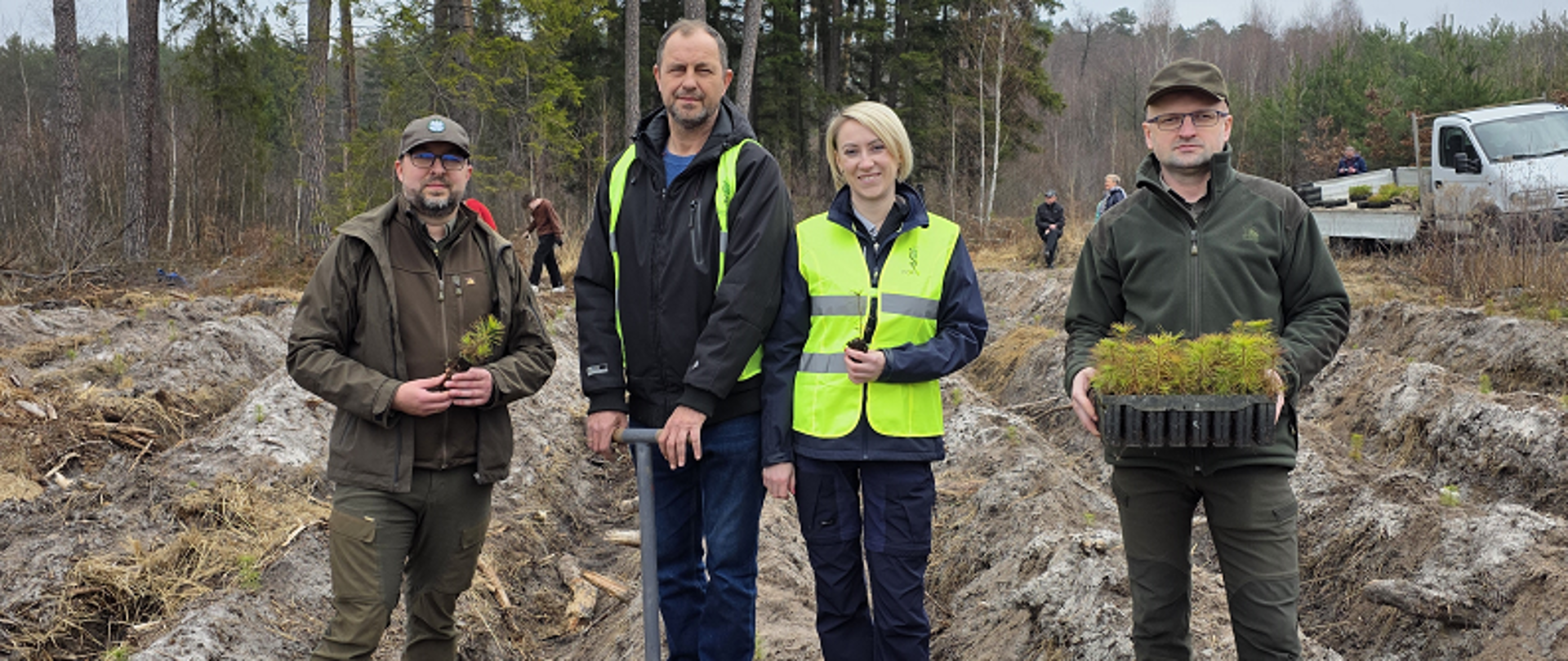 Cztery osoby stoją na rozkopanym polu. Mężczyzna po prawej stronie trzyma w dłoniach pojemnik z sadzonkami. Mężczyzna w środku wbija w ziemię kostur sadzarski. W tle las i samochód cieżarowy. 