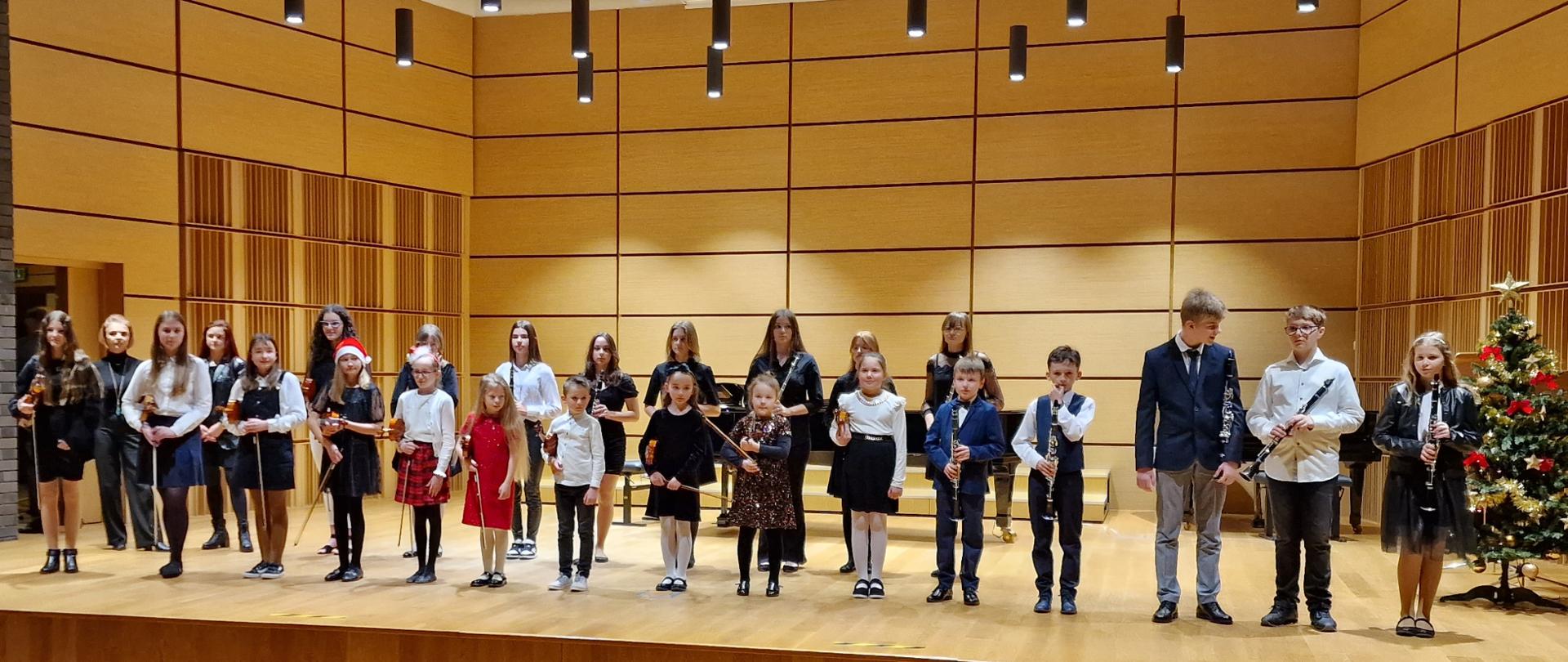Zdjęcie przedstawia uczniów klas klarnetu, fletu i skrzypiec oraz nauczycieli ustawionych w dwóch rzędach. Na dole widnieje widownia sali koncertowej. Po prawej stronie na scenie ozdobiona choinka.