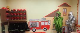 Na zdjęciu ubrania strażackie i hełmy dla dzieci oraz ubranie gazoszczelne i żaroochronne. Na ścianie namalowany samochód strażacki i dom z widocznym pożarem w oknie. 