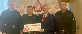 Trzech strażaków i prezes klubu „Jednostka HDK” pozują na tle ścianki z logo klubu. W rękach trzymaja tabliczkę z napisem z krwią na ratunek z klubem jednostkaHDK