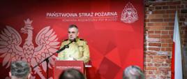 Na zdjęciu widać mężczyznę w mundurze Państwowej Straży Pożarnej stojącego za mównicą i wygłaszającego przemówienie. Tło stanowi czerwony baner z godłem Polski oraz logo Państwowej Straży Pożarnej, a także napis „Komenda Wojewódzka PSP w Łodzi”. W tle znajduje się ceglana ściana i flaga Polski. W pierwszym planie widoczne są plecy kilku słuchaczy, niektórzy z nich również noszą mundury.