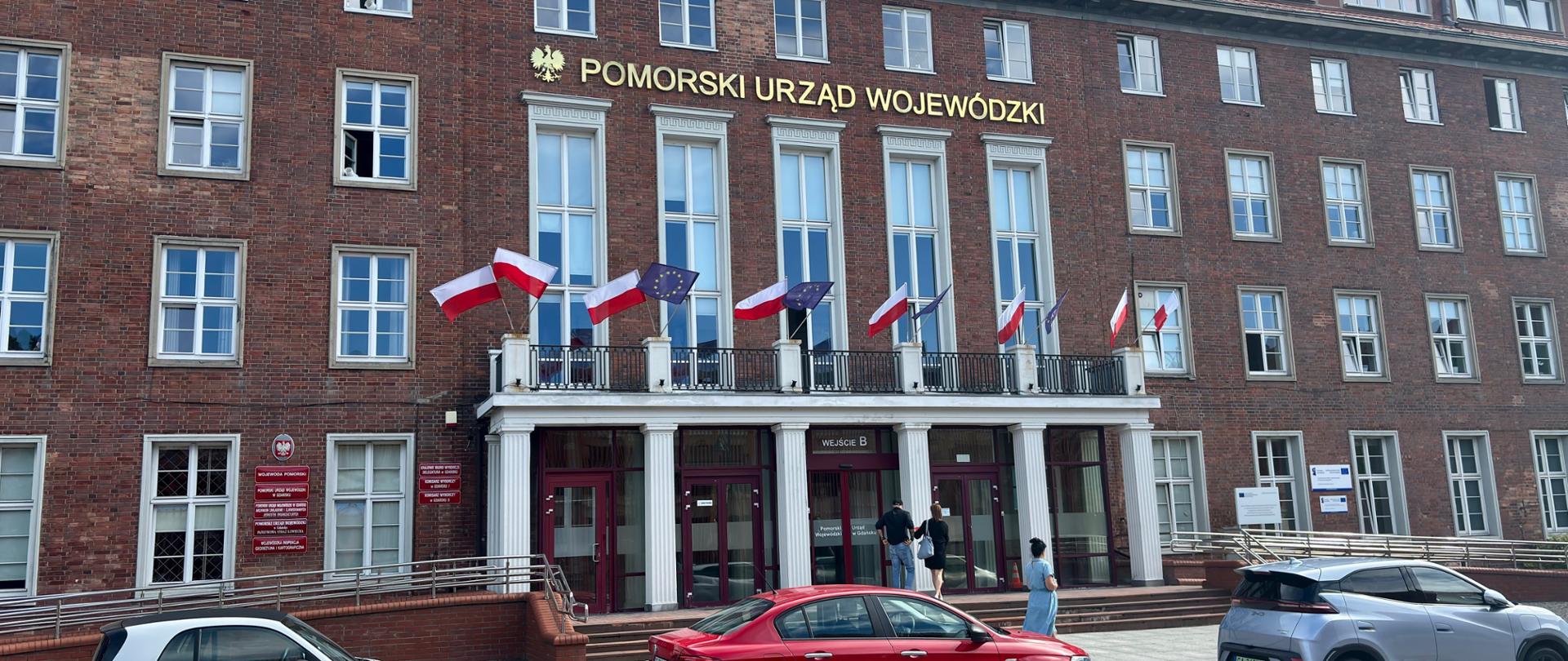 Budynek Pomorskiego Urzędu Wojewódzkiego, z brązowej cegły, przy wejściu białe filary i flagi Polski i Unii Europejskiej