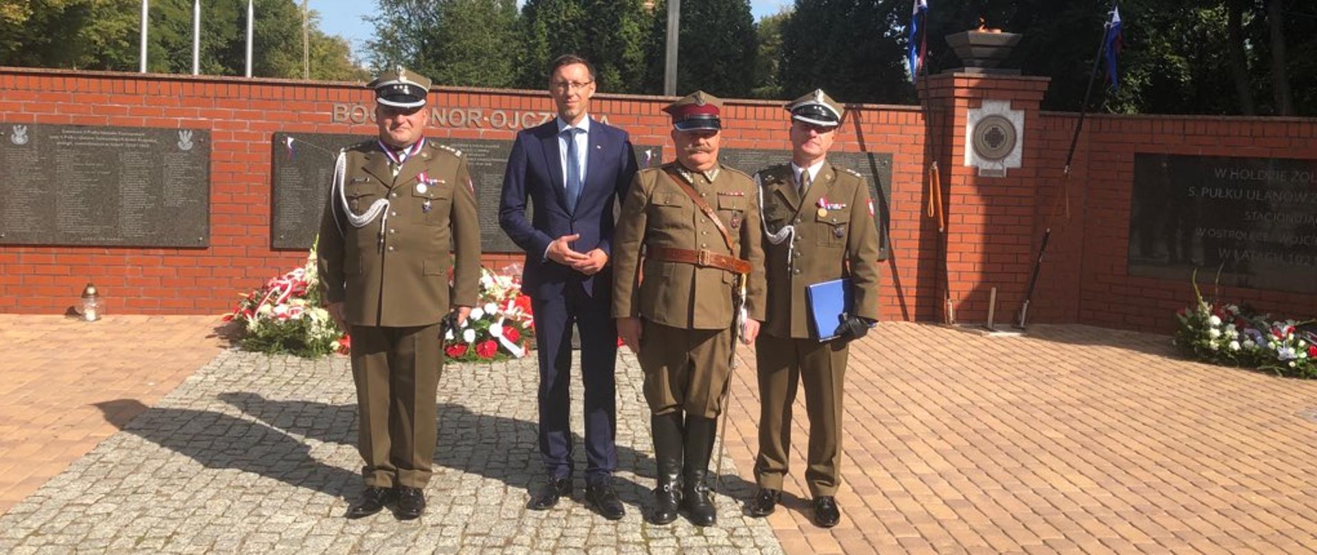 Na zdjęciu widać czterech mężczyzn: jeden ubrany w granatowy garnitur stoi pomiędzy trzema wojskowymi. Mężczyźni stoją przed pomnikiem
