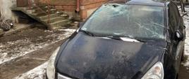 Na zdjęciu samochód osobowy barwy czarnej z uszkodzoną przednią szybą czołową, w tle budynek mieszkalny z uszkodzonym pokryciem dachowym na który przykryto plandeką zabezpieczającą, niebo pochmurne z przejaśnieniami, za budynkiem widoczny samochód ciężarowy ochotniczej straży pożarnej