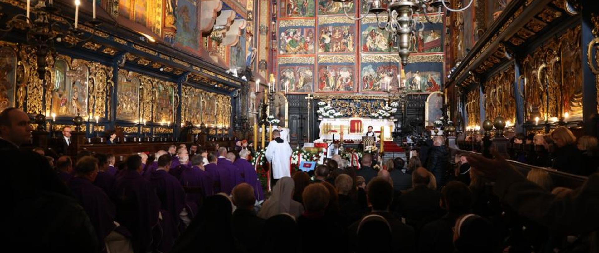 Widok z tyłu na wielką kościelną salę, ściany zdobione kolorowymi obrazami, na ławkach siedzi dużo ludzi, z przodu ołtarz.