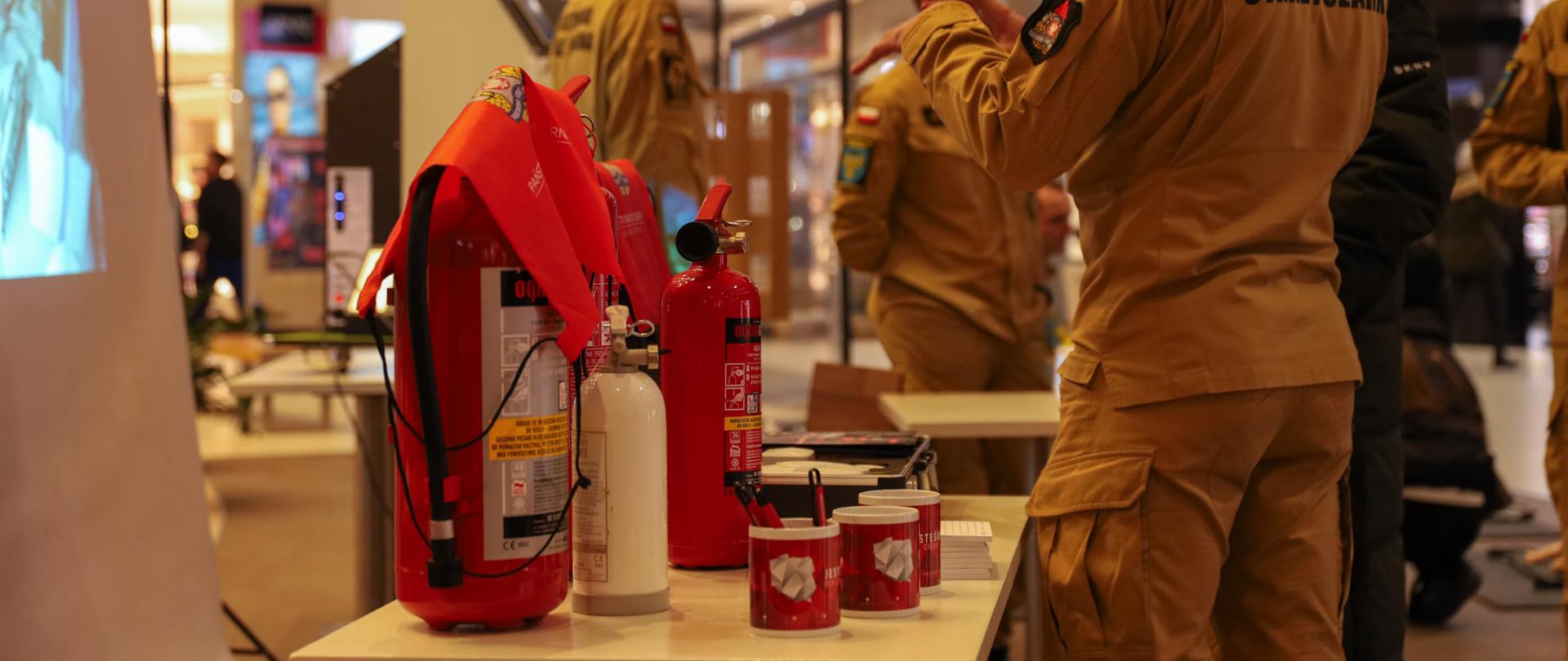 Strażak doradza w wyborze czujki do domu. W tle gaśnice i gadżety promocyjne.