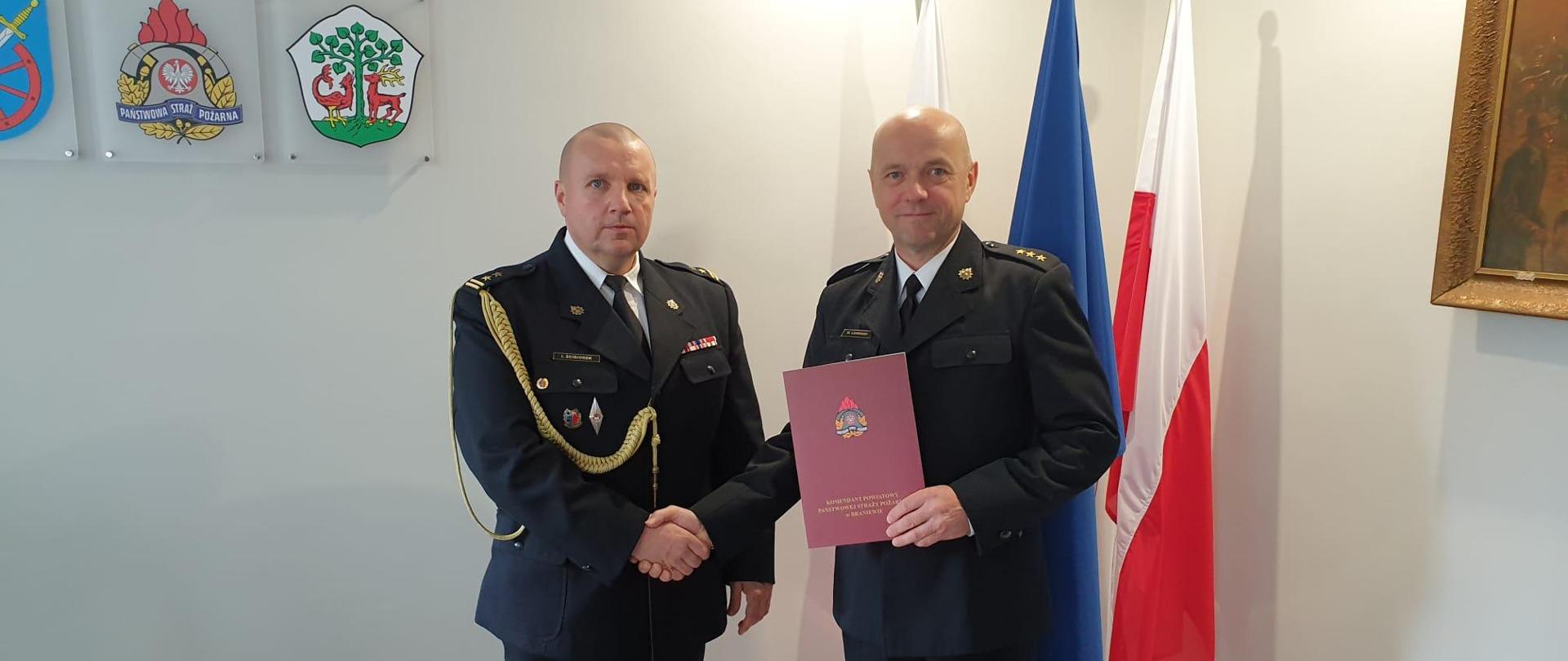 NA zdjęciu w gabinecie, komendant powiatowy wręcza dokument dla dowódcy jednostki. W tle flaga państwowa a na ścianie godło państwowe, logo PSP, powiatu braniewskiego i miasta Braniewa
