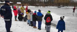 Trzech funkcjonariuszy Państwowej Straży Pożarnej pokazuje grupie dzieci jak należy zachować się w chwili, gdy pod kimś zarwie się lód. Jeden ze strażaków zmierza po lodzie w kierunku przerębla, a dwóch go asekuruje. Stojące na brzegu dzieci wraz z policjantem obserwują pokaz. Wszyscy znajdują się nad stawem w Miejskim Ośrodku Sportu i Rekreacji w Rypinie.