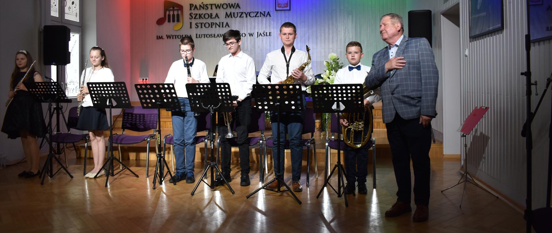 Zespół instrumentalny w składzie flet, trąbka, klarnet, saksofon, sakshorn. Obok nich dyrygent w niebieskiej marynarce. Wszyscy czekają na ukłon.