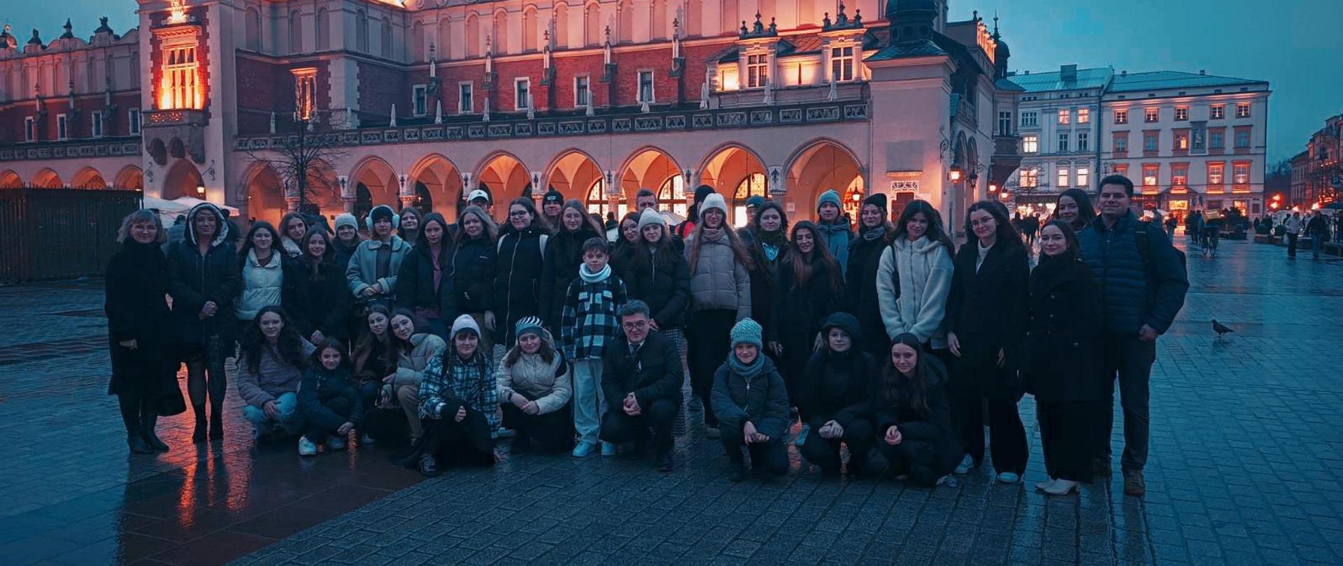 uczniowie naszej szkoły wraz z nauczycielami wieczorem na krakowskim rynku na zdjęciu grupowym przed budynkiem sukiennic