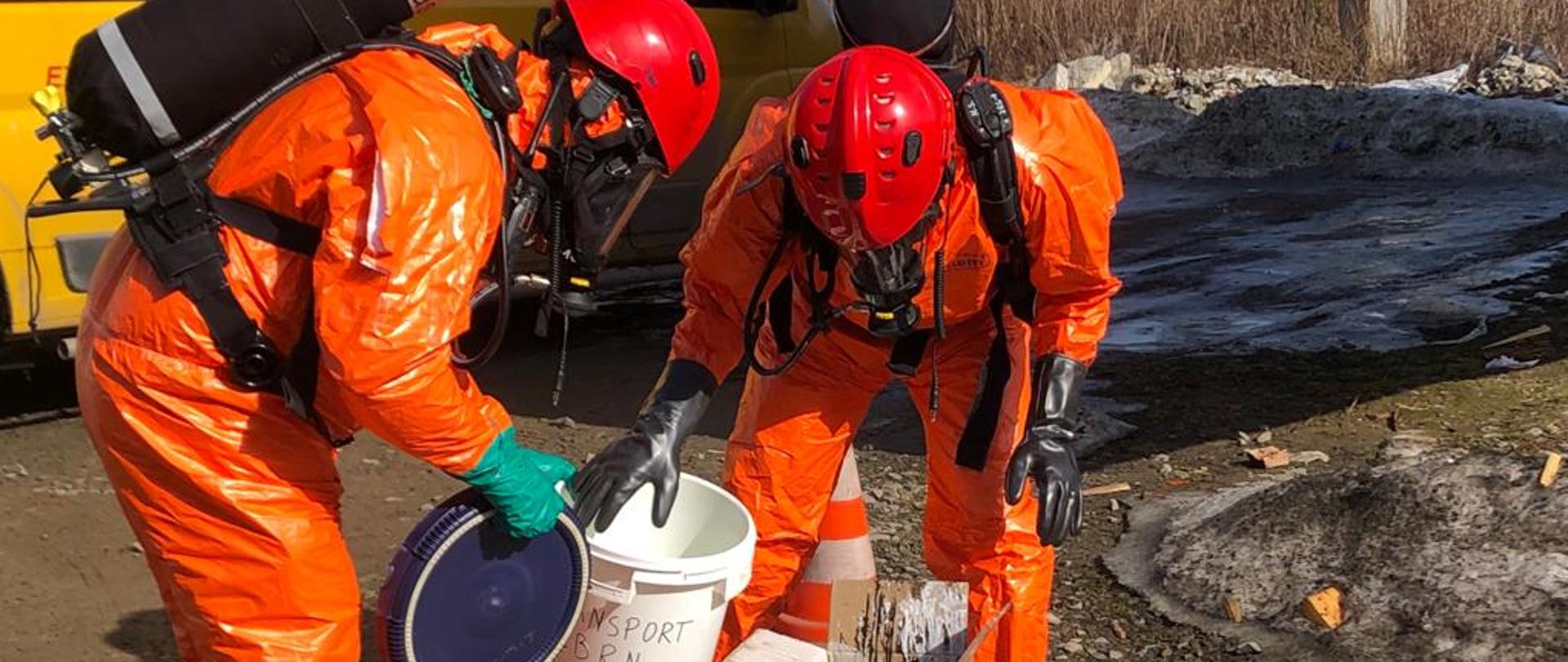 Dwóch strażaków w pomarańczowych kombinezonach ochronnych prowadzi działania w wyznaczonym sektorze.