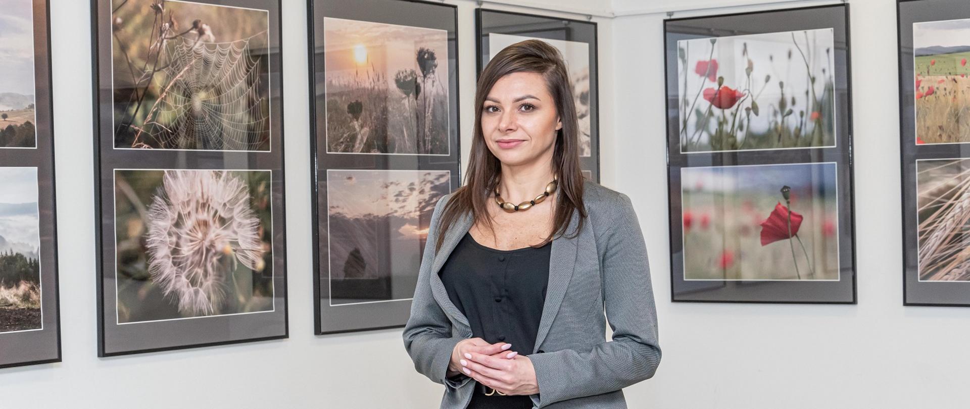 Na zdjęciu autorka wystawy, która stoi na tle ściany gdzie wiszą zdjęcia jej autorstwa. 