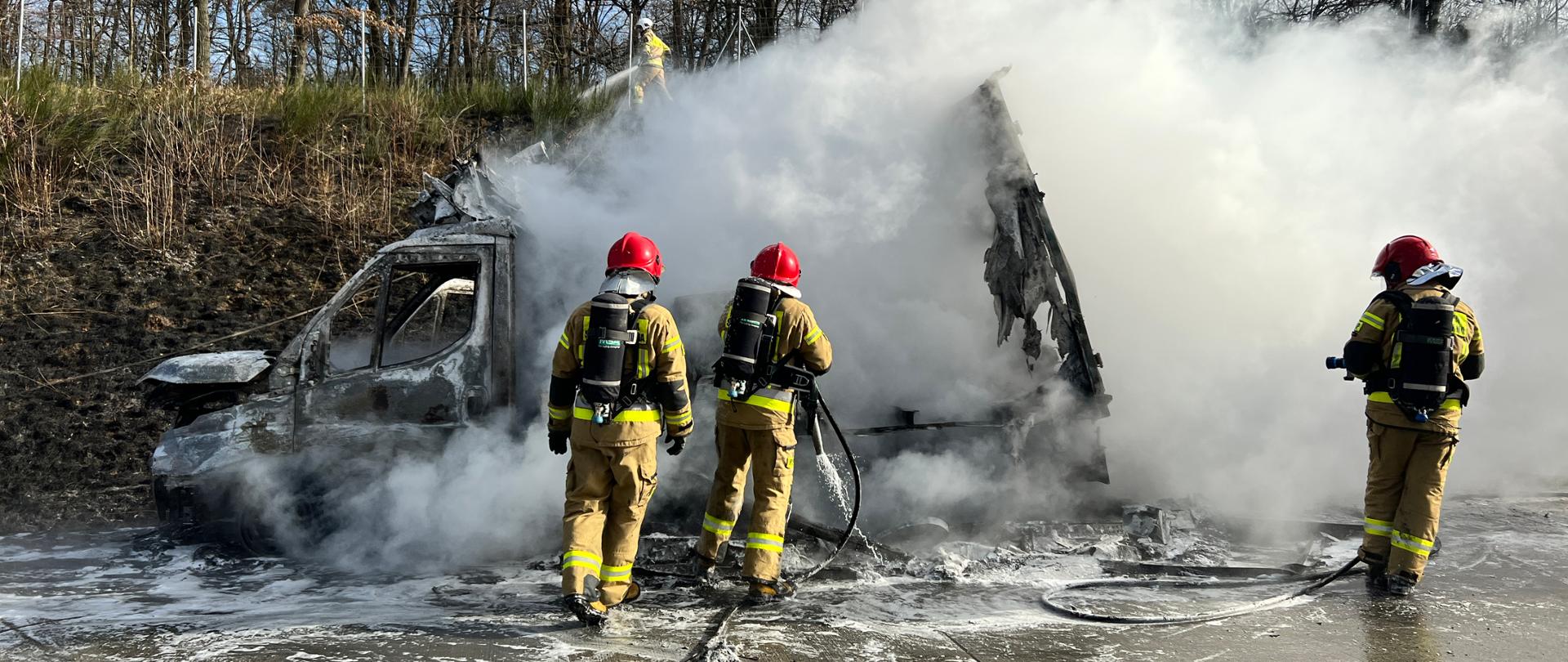 strażacy gaszący pojazd typu bus, widoczne zadymienie, działania prowadzone na autostradzie, w tle skarpa z zadrzewieniem