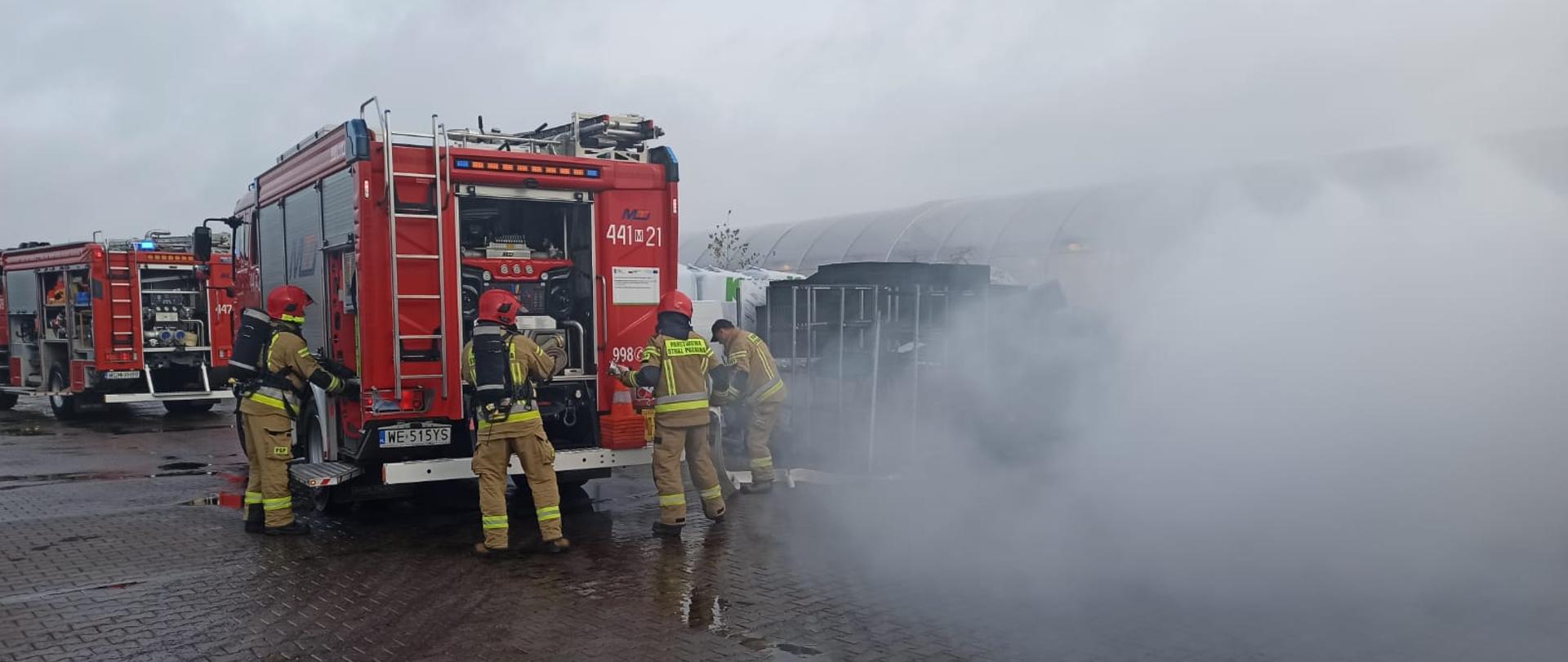 Strażacy wyjmują sprzęt z samochodu pożarniczego i przygotowują się do rozwijania węży w tle widać pozorowane zadymienie. 