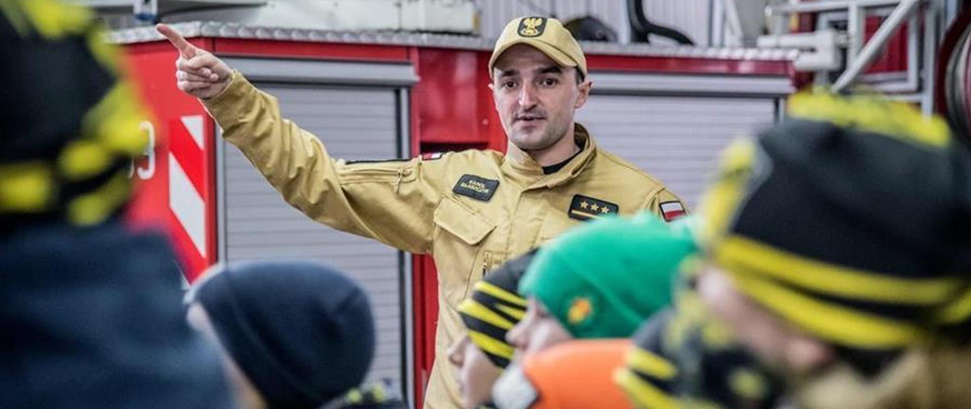 Strażak w ubiorze służbowym, który ręką pokazuje znajdującym się dzieciom na garażu samochody ratowniczo-gaśnicze.