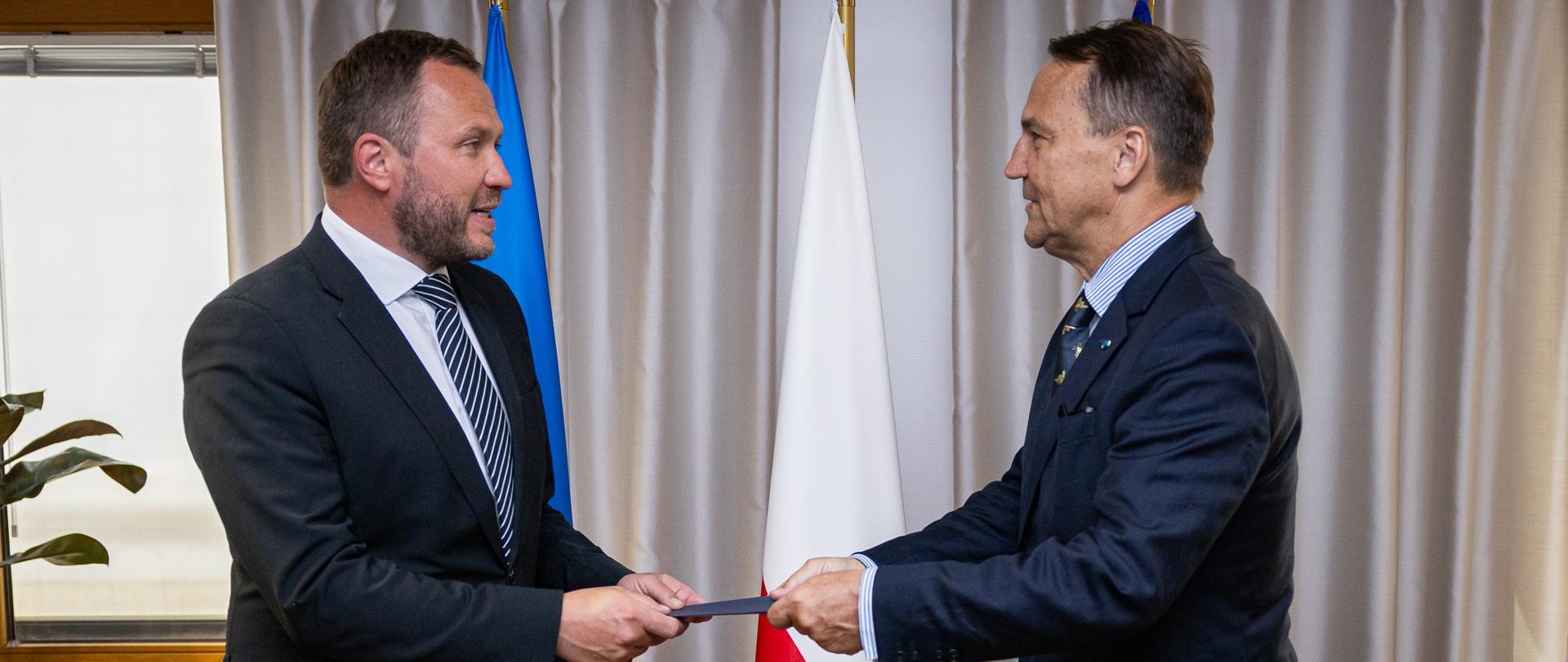 1 lipca Polska przejmuje przewodnictwo w Radzie Państw Morza Bałtyckiego, na zdjęciu minister spraw zagranicznych Estonii przekazuje teczkę -symbolicznie prezydencję w Radzie- ministrowi spraw zagranicznych RP. Mężczyźni w garniturach stoją na tle flagi Estonii, Polski i Unii Europejskiej.