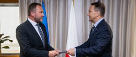 1 lipca Polska przejmuje przewodnictwo w Radzie Państw Morza Bałtyckiego, na zdjęciu minister spraw zagranicznych Estonii przekazuje teczkę -symbolicznie prezydencję w Radzie- ministrowi spraw zagranicznych RP. Mężczyźni w garniturach stoją na tle flagi Estonii, Polski i Unii Europejskiej.