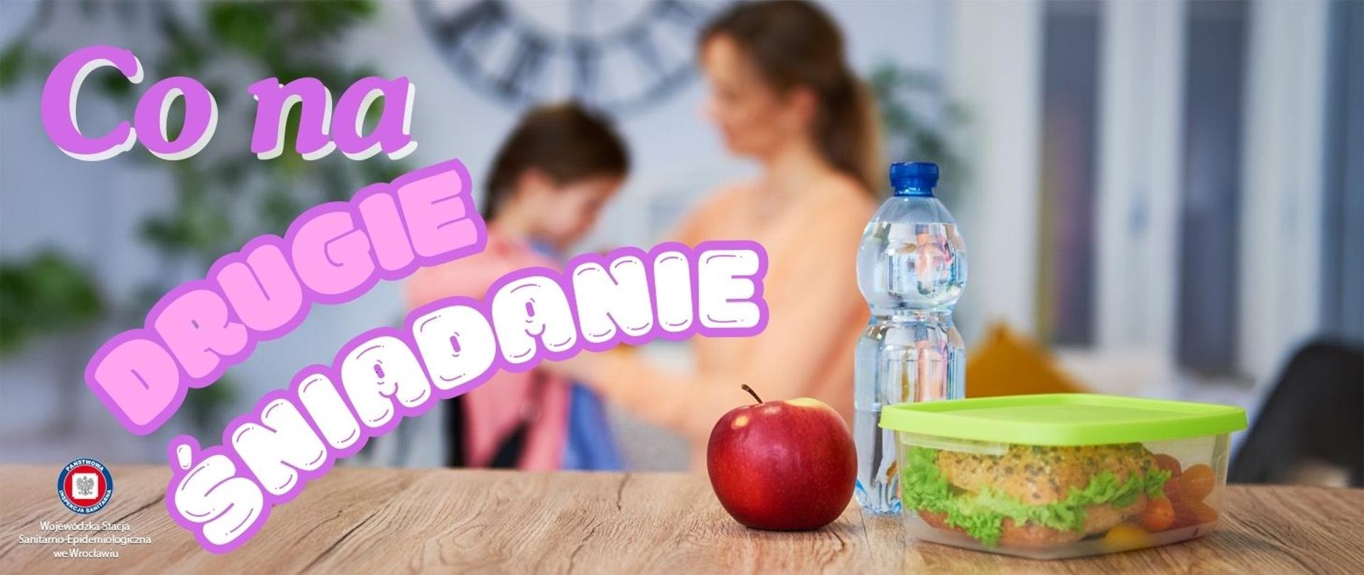 Mama przygotowująca córkę do szkoły. Na pierwszym planie na stole lunchbox ze zdrową kanapką butelka wody oraz czerwone jabłko.