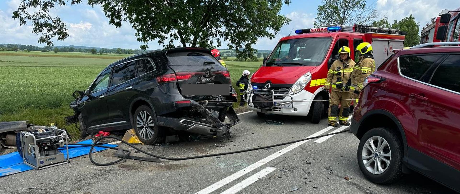Obraz przedstawia rozbity pojazd. W tle samochód pożarniczy i strażacy. Pora dzienna.