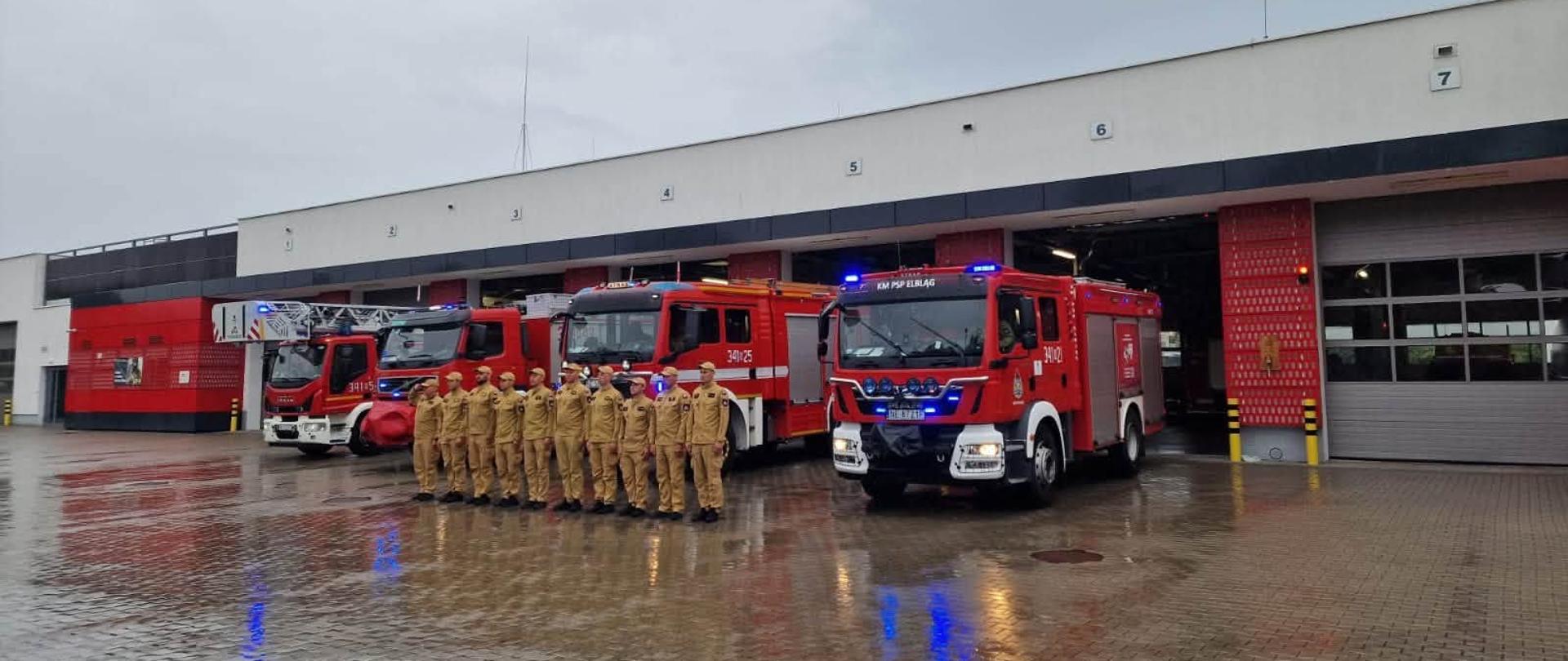 Na zdjęciu widoczna jest uroczysta zbiórka strażaków przed budynkiem jednostki. Funkcjonariusze stoją w równym szyku na placu manewrowym, w pełnym umundurowaniu służbowym. W tle ustawione są wozy pożarnicze z włączonymi sygnałami świetlnymi, podkreślając podniosły charakter wydarzenia.