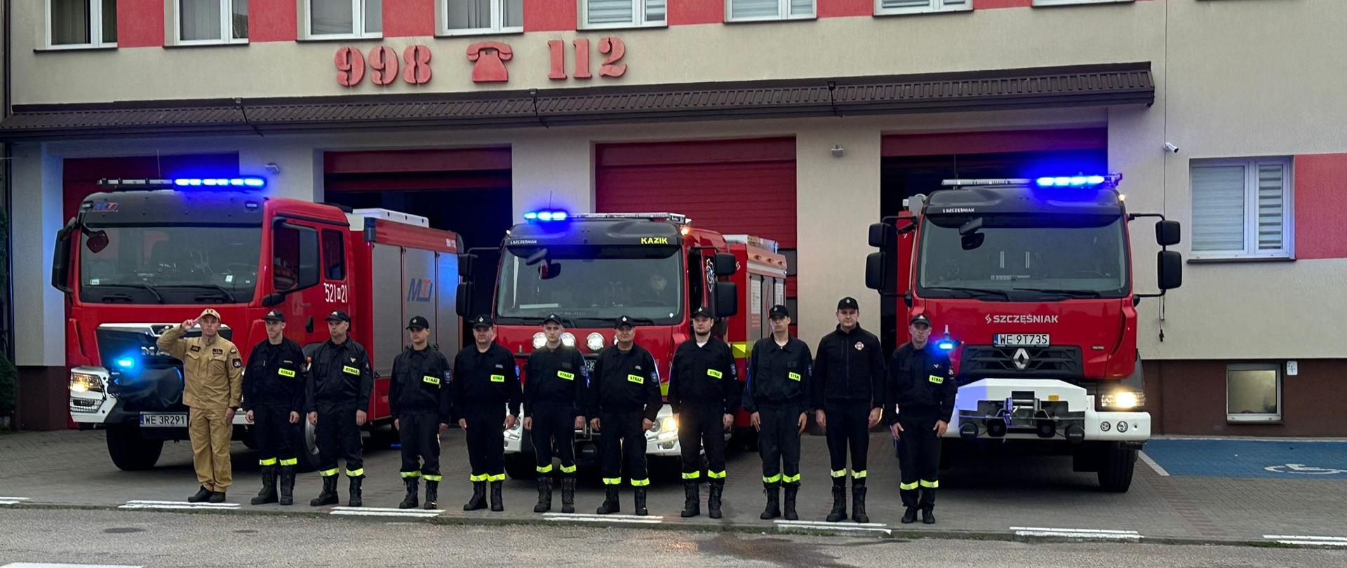 Grupa strażaków w galowym ustawieniu stoi w szeregu przed budynkiem Państwowej Straży Pożarnej. Za nimi zaparkowane są trzy czerwone wozy strażackie z włączonymi sygnałami świetlnymi. Na budynku widoczny jest napis „Państwowa Straż Pożarna” oraz numery alarmowe 998 i 112. Jeden ze strażaków w beżowym mundurze oddaje salut. Na twarzach obecnych powaga — zdjęcie wykonane w hołdzie poległemu druhowi.