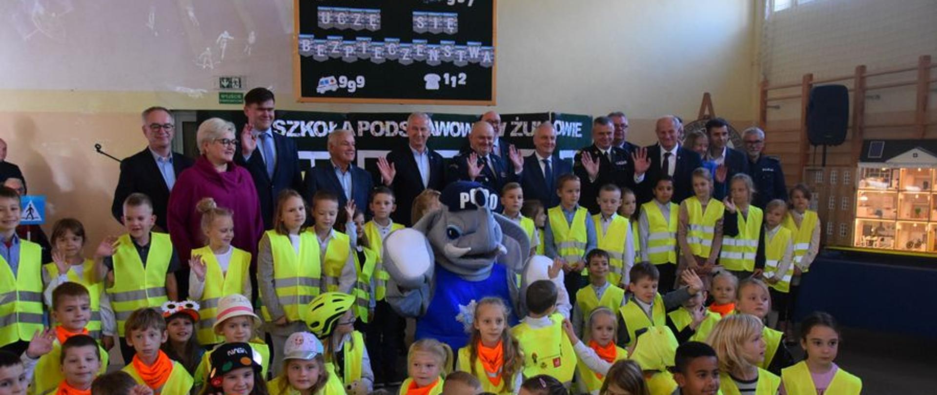 Dzieci ubrane w kamizelki odblaskowe oraz zaproszeni goście stoją obok siebie.