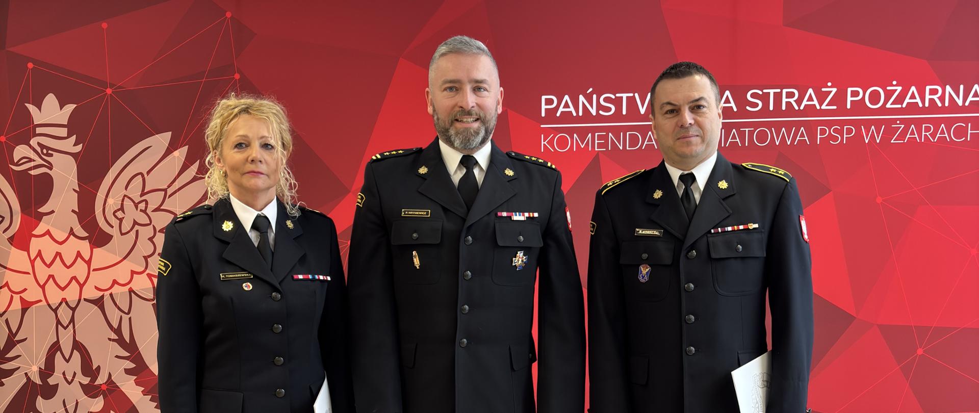 Strażacy otrzymujący nagrodę z Komendantem w czarnych mundurach mundurach wyjściowych, w tle czerwony baner Państwowej Straży Pożarnej