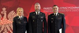 Strażacy otrzymujący nagrodę z Komendantem w czarnych mundurach mundurach wyjściowych, w tle czerwony baner Państwowej Straży Pożarnej