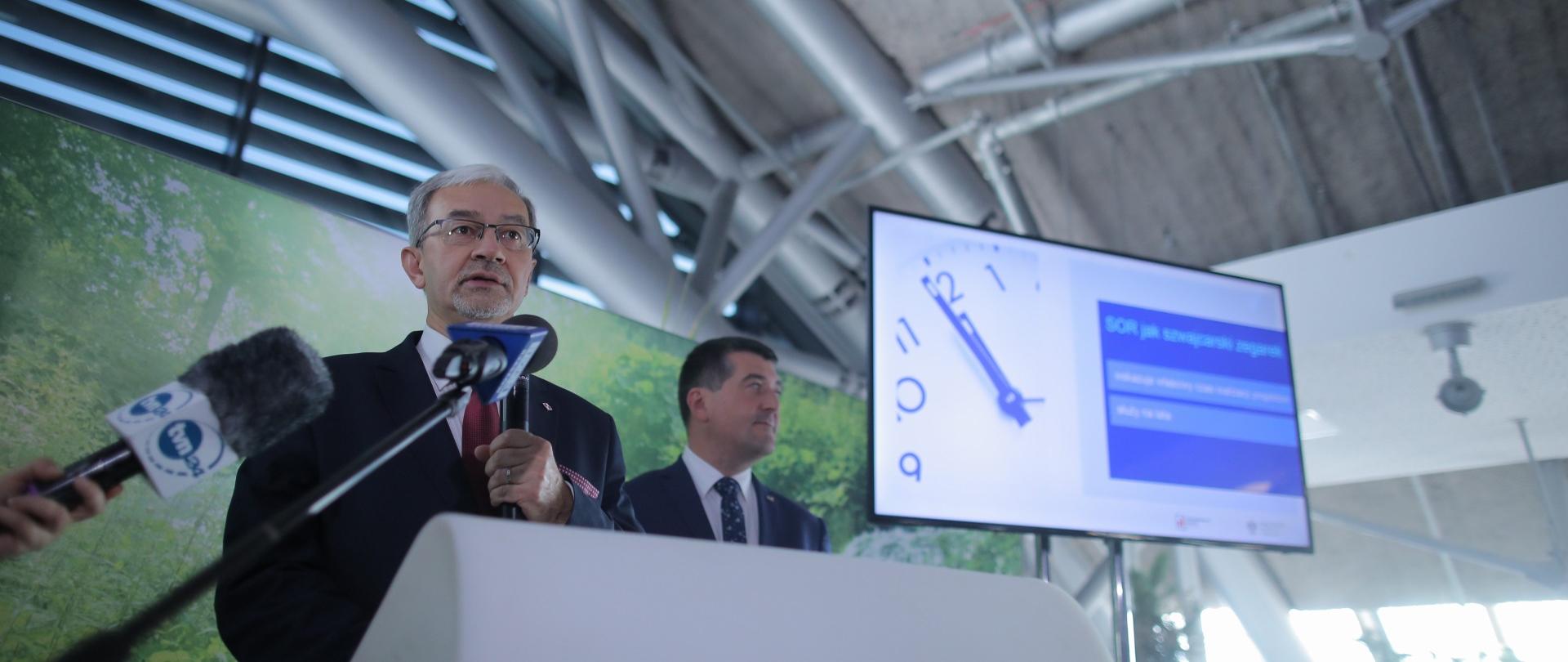 Minister Kwieciński stoi przy mównicy, przed nim znajdują się mikrofony dziennikarskie, za nim wyświetlana jest grafika z napisem "SOR jak szwajcarski zegarek", na grafice jest zdjęcie zegara. 